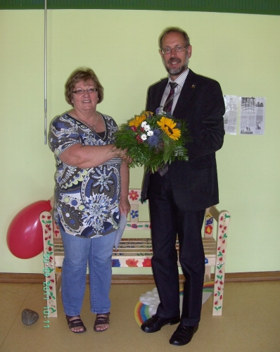 Bürgermeister Miola verabschiedet Frau Kunig mit einem Blumenstrauß. Im Hintergrund ist die selbstbemalte Bank der Kindergartenkinder zu sehen.
